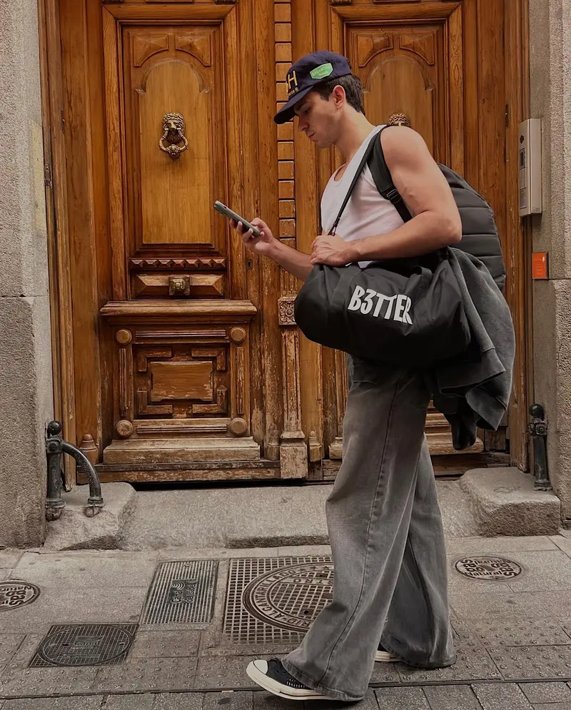 Adrià Cruz de perfil, con gorra y el móvil, ante un portal de madera; lleva una bolsa de B3TTER.